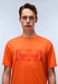 Oranž puuvillane T-särk ümmarguse kaelusega ja lühikeste varrukatega. Ees on pleekinud grafika, sealhulgas maakera ja tekstilogo.