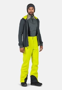 Pantaloni impermeabili gialli con accenti neri, abbinati a un top grigio a maniche lunghe e un cappello lavorato a maglia scuro. Presentano spallacci regolabili e una tasca con zip.