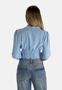 Helles blaues, kurz geschnittenes Blusenshirt mit lange, puffigen Ärmeln und einem gerüschten Bund. Getragen mit hoch taillierten Jeans aus Denim. Glatte Textur, lässiges Design.
