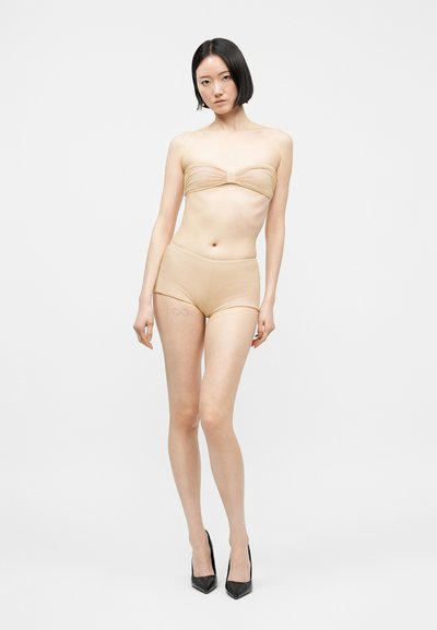 Femme aux cheveux courts noirs portant un haut bandeau beige et un short taille haute, debout en talons noirs pointus sur un fond blanc.