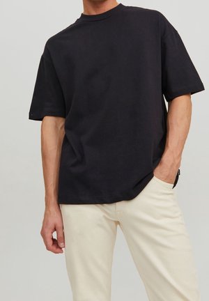 T-shirt en coton noir à manches courtes et col rond, associé à un pantalon tapered de couleur claire. Le tissu semble lisse et sans texture.