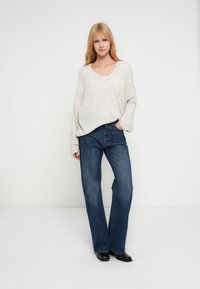 Camisola cinzento claro em malha com decote em V e corte amplo, combinada com jeans azul escuro de perna larga. O conjunto apresenta um estilo descontraído e casual.