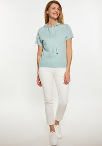 Felpa verde menta a maniche corte con cappuccio e cordino, indossata con pantaloni bianchi slim-fit e sneakers bianche. Texture liscia, design casual.