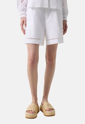 Kobieta w białych szortach z ozdobnymi wycięciami przy krawędziach, białym topie oraz beżowych sandałach na platformie z logo "B".