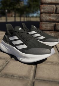 Le scarpe da corsa grigio scuro presentano una parte superiore texturizzata, un design a tre strisce bianche e una suola bianca ammortizzata, posizionate su una superficie pavimentata.