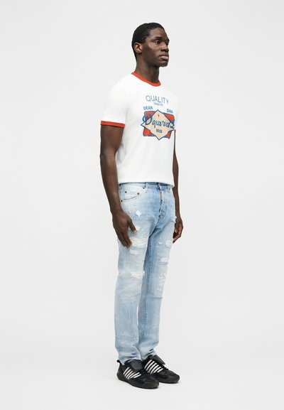 Homme portant un t-shirt blanc à motif avec bordure rouge, un jean bleu clair déchiré et des baskets noires, debout devant un fond uni.