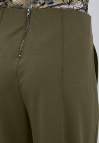 Pantalons vert olive en tissu lisse avec une fermeture à glissière sur le côté et un design épuré, présentant des lignes nettes et un minimum de détails.