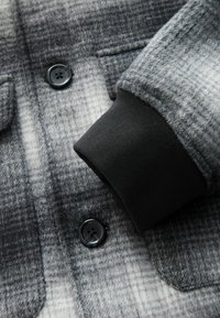 Gråternet skjorte lavet af struktureret uld, med sort knap, tydelig lomme og ribbet sort manchet detalje.