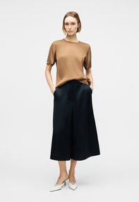 Une femme se tient debout, les mains dans les poches, portant un haut marron à manches courtes, un pantalon noir large et court, ainsi que des talons blancs pointus sur un fond blanc.