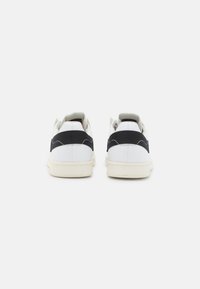 Bílé tenisky s černými textilními akcenty, nízký design, gumová podrážka a texturované prošití na zadní straně.