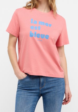 Femme portant un t-shirt rose vif avec un texte bleu "la mer est bleue" et un jean bleu, main dans la poche, sur fond clair uni.