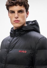 Chaqueta acolchada negra con un acabado brillante, cierre frontal con cremallera y capucha ajustable. Presenta el logo rojo "HUGO" en la zona del pecho izquierdo.