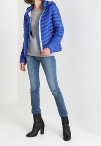 Blå polstret jakke med hætte, hvid lynlås og logo på armen, parret med en grå strikket trøje, blå jeans og sorte hælede støvler.