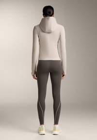 Ženska stoji obrnjena stran, oblečena v svetlo bež kapucar jakno, temno sive legice z odsevnim šivom in bele superge z rumenimi poudarki.