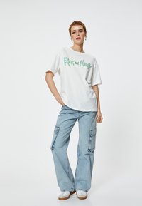 T-shirt bianca con grafica verde "Rick and Morty", abbinata a pantaloni cargo azzurri chiari con multiple tasche e vestibilità rilassata.