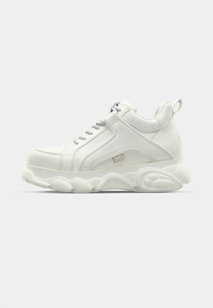 Tênis atléticos brancos com parte superior em couro liso, sola arredondada e detalhes texturizados. Possui cadarços e uma alça na parte de trás.