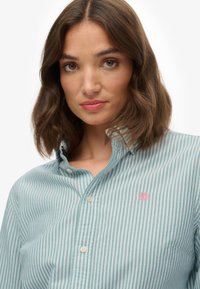 Turkusowa koszula w pasy z krótkim rękawem, z kołnierzykiem, z małym różowym logo na lewym piersi oraz białymi guzikami z przodu.