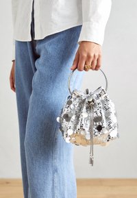 Silverfärgad paljettsäck med ett cirkulärt metallhandtag, som visar en dragskoöppning och reflexiva detaljer. Bärs med jeansbyxor.