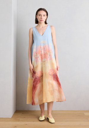 Femme debout contre un mur uni, portant une robe sans manches à col en V avec des motifs abstraits de paysages pastel et des chaussures jaunes sans lacets.