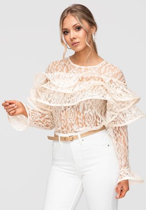 Jeune femme portant un chemisier en dentelle blanche transparente avec des volants superposés, un pantalon blanc taille haute et une ceinture beige, posant devant un fond uni.