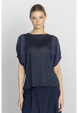 Kobieta z włosami związanymi do tyłu, ubrana w luźną granatową bluzkę z bufkami i dopasowaną spódnicę, stojąca na jednolitym jasnym tle.