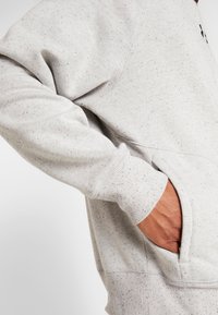 Felpa grigia chiara con texture a macchie, caratterizzata da polsini a coste e una tasca laterale. Il tessuto appare morbido e resistente.