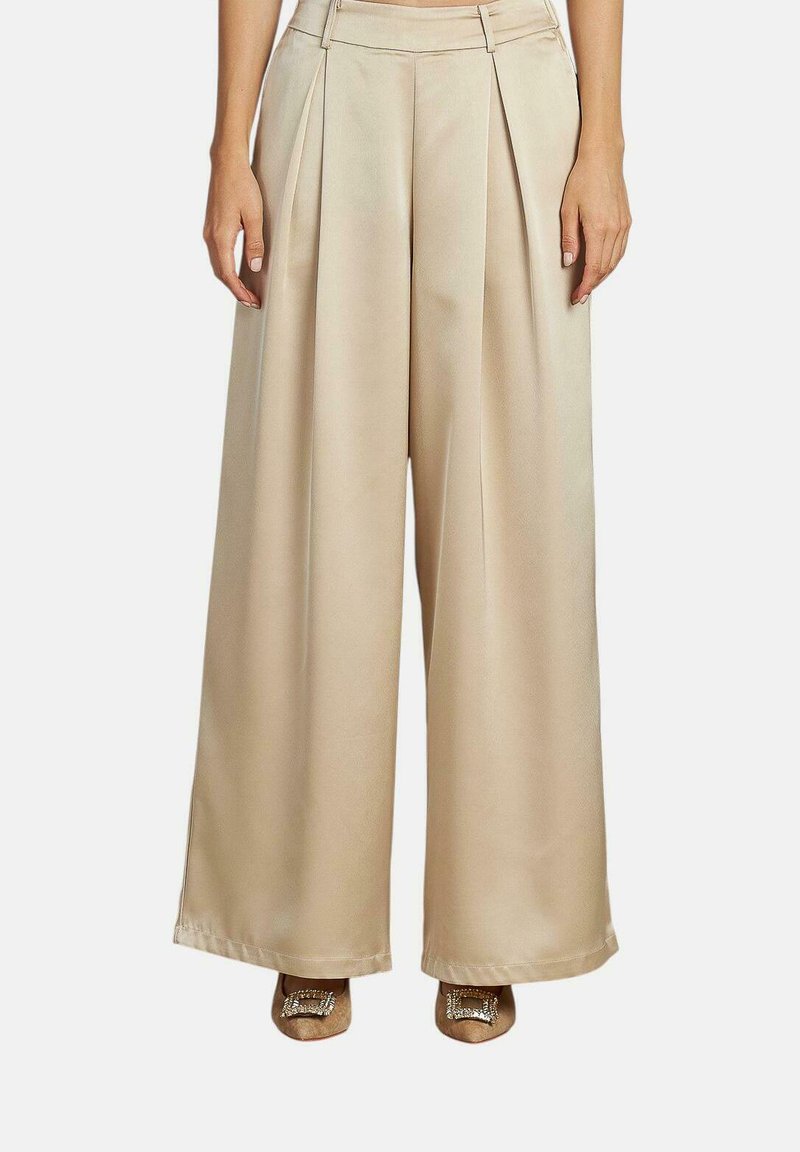 Pantalons beiges à jambes larges avec plis, en tissu lisse, présentant une taille haute et une coupe structurée, accompagnés de chaussures à bout pointu.