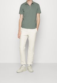 Polo en vert gris avec un col zippé, associé à un pantalon blanc et des baskets claires. Le tissu semble doux et a une coupe décontractée.