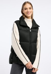 Zwarte puffer vest met hoge kraag, ritssluiting aan de voorkant en knopen aan de zijkant. Heeft een textuur wit ribbeltrui eronder.
