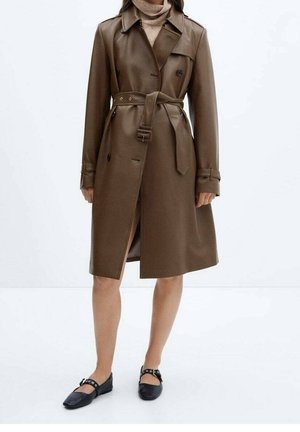 Vrouw draagt een bruine leren trenchcoat met riem over een beige coltrui, gecombineerd met zwarte Mary Jane platte schoenen.
