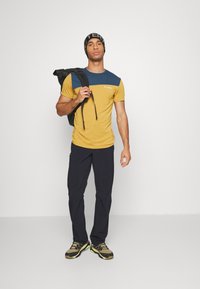 Tenue de plein air composée d'une chemise à manches courtes jaune et bleue, de pantalons foncés, et de chaussures de randonnée, accompagnée d'un sac à dos noir et d'un bonnet.