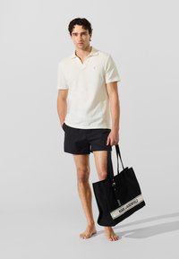 Camiseta polo de color crema claro con cuello, bañador negro y un gran bolso tote negro con una banda de logo blanca, llevado por una persona.