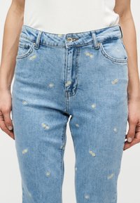 Ανοιχτό μπλε τζιν με μικρά κεντημένα λευκά και κίτρινα σχέδια μαργαρίτας κατανεμημένα σε όλο το ύφασμα, φορεμένα με λευκό πουκάμισο.