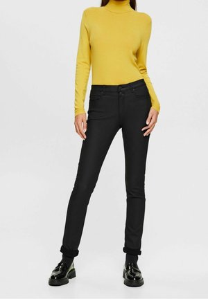 Nainen, jolla on tiukka keltainen pooloneule, mustat kapeat farkut, joiden lahkeet on käännetty nilkan kohdalla, ja mustat kiiltävät loafersit, seisoo yksivärisen taustan edessä.