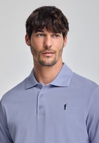 Polo shirt di colore lilla chiaro con un tessuto texturizzato, tre bottoni e un logo ricamato di un golfista in nero sul lato sinistro del petto.