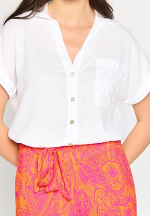 Kobieta ubrana w krótką białą koszulę na guziki, włożoną w wysokowatą spódnicę w różowo-pomarańczowy wzór, z wiązaniem z przodu.