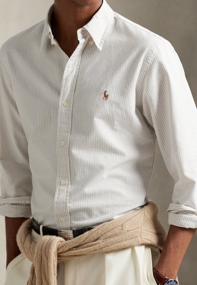 Camisa às riscas em bege claro e branco com um pequeno logótipo bordado. Suéter de malha castanho claro amarrado na cintura. Apresenta colarinho clássico.