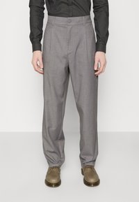 Lichtgrijze geplooide broek van gestructureerde stof, voorzien van een knoopsluiting en een losse pasvorm, gecombineerd met bruine schoenen.