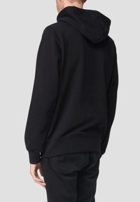 Persona che indossa un hoodie nero e pantaloni neri, vista da dietro su uno sfondo grigio uniforme.