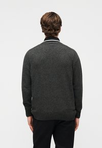 Άνδρας που φαίνεται από πίσω, φοράει σκουρόχρωμο γκρι πλεκτό πουλόβερ πάνω από μαύρο πουκάμισο με γιακά και μαύρο παντελόνι, μπροστά σε ένα απλό λευκό φόντο.