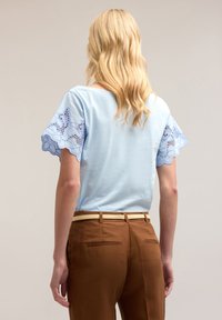 Lichtblauw top met decoratieve kantmouwen. Bruine broek met een beige riem, voorzien van achterzakken en een getailleerde pasvorm. Gladde stoftextuur.