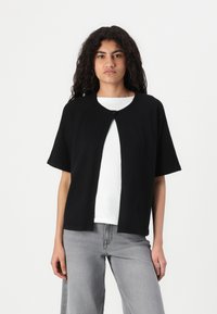 Black short-sleeve cardigan, open front with a single button closure, soft fabric, casual design, paired with a white shirt and gray pants.