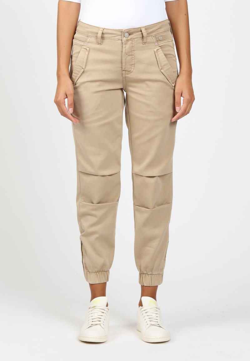 Femme portant un pantalon beige fuselé avec poches avant et poignets élastiques, associé à des baskets blanches sur un fond uni.