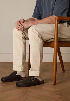Sabots en cuir marron foncé avec un large bandeau et un détail de boucle, portés sur des chaussettes gris clair, associés à un pantalon de couleur crème et une chemise bleue.