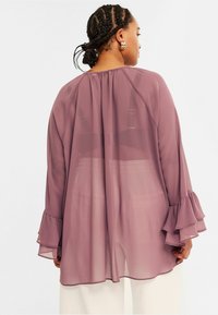 Lila chiffongblus med långa ärmar, volangärmar och en lös passform. Baksidan har en något genomskinlig design med en rundad nederkant.