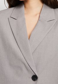 Harmaa muotoiltu bleiseri, jossa on lovipanta, kevyesti teksturoitu kangas, yksittäinen musta nappilukitus ja hienovaraiset ompeluyksityiskohdat.