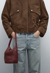 Bruin suède cropped jack met twee zakken, gecombineerd met lichtblauwe jeans. Heeft een kleine bordeauxrode geweven tas die met de linkerhand wordt vastgehouden.