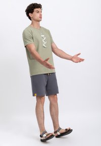 Olijfgroene t-shirt met een grafisch ontwerp, gecombineerd met donkergrijze shorts met een gele accenten. Model draagt grijze sandalen.