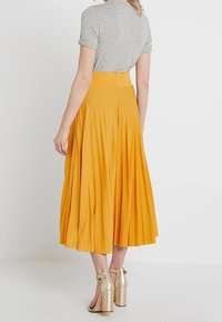 Falda midi amarilla con plisados y cintura alta, combinada con una blusa gris de manga corta y sandalias doradas de tacón. Tejido suave con un diseño fluido.