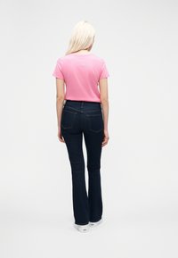 Ξανθιά γυναίκα με πλάτη, φοράει ροζ κοντομάνικη μπλούζα, σκούρο μπλε τζιν παντελόνι bootcut και λευκά αθλητικά παπούτσια σε απλό φόντο.